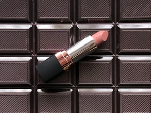 A pink lipstick rests on a background featuring a pattern resembling a chocolate bar. The lipstick has a sleek silver and black casing with a rose gold accent. The chocolate bar background has distinct, glossy square patterns.