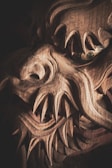 A detailed close-up of a wooden carving featuring intricate and sharp features, resembling a mythical creature or dragon with fierce expressions. The complex wood grain and elaborate carving techniques are prominently visible, highlighting skillful craftsmanship.