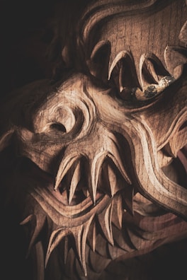 A detailed close-up of a wooden carving featuring intricate and sharp features, resembling a mythical creature or dragon with fierce expressions. The complex wood grain and elaborate carving techniques are prominently visible, highlighting skillful craftsmanship.