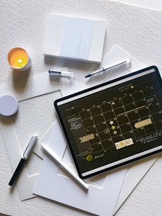 An arrangement featuring a planner, a lit candle, several pens, a digital tablet displaying a calendar, and some white sheets of paper. The planner is closed and has a minimalist design, while the digital tablet is in landscape mode, showing a detailed monthly calendar. The pen lids are off, and the candle is in a small holder, casting a soft light.