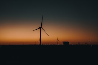 Wind turbines silhouetted against a sunset, with the sky transitioning from a warm orange near the horizon to a darker blue as it extends upward. Several turbines are visible on the horizon line, along with other indistinct structures.