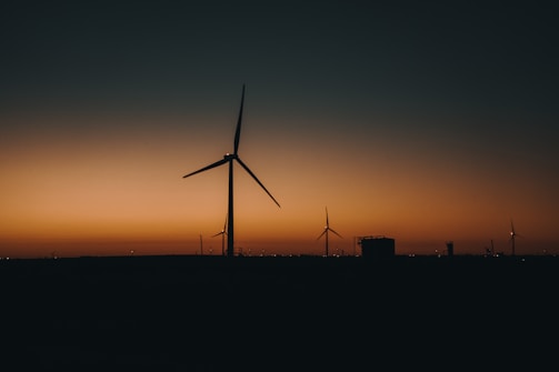 Wind turbines silhouetted against a sunset, with the sky transitioning from a warm orange near the horizon to a darker blue as it extends upward. Several turbines are visible on the horizon line, along with other indistinct structures.