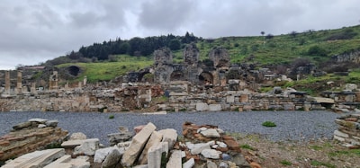 Ancient ruins are scattered across a hilly landscape, with remnants of stone arches and columns visible amidst the rubble. The stones and structures show signs of significant wear and age. The background features a lush hillside covered with vegetation and a cloudy sky overhead, contributing to the historical ambience.