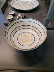 A collection of intricately designed ceramic dishes featuring Arabic calligraphy, with a large shallow bowl prominently displayed in the foreground. The bowl is adorned with blue and gold script encircling the interior, while smaller cups and vessels sit elevated in the background.