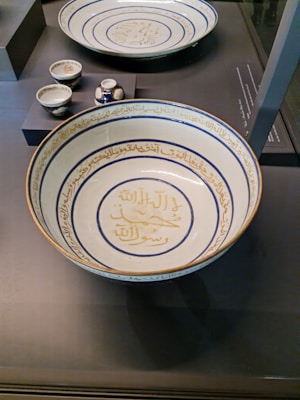 A collection of intricately designed ceramic dishes featuring Arabic calligraphy, with a large shallow bowl prominently displayed in the foreground. The bowl is adorned with blue and gold script encircling the interior, while smaller cups and vessels sit elevated in the background.