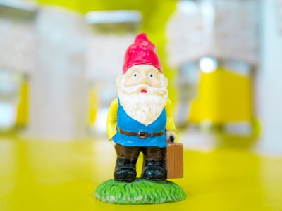 A small figurine of a gnome with a white beard and red hat is standing on a grassy base. The gnome is dressed in a blue shirt and brown pants, holding a brown suitcase in its right hand. The background is blurred, featuring a mix of yellow and white hues.