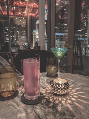 A dimly lit bar setting featuring a marble table with various drinks and a decorative candle holder casting intricate light patterns. A pink cocktail sits in a tall glass, accompanied by a green martini glass with a garnish. The background shows a window view of city buildings and lights against a nighttime sky.