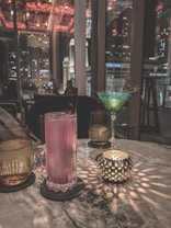 A dimly lit bar setting featuring a marble table with various drinks and a decorative candle holder casting intricate light patterns. A pink cocktail sits in a tall glass, accompanied by a green martini glass with a garnish. The background shows a window view of city buildings and lights against a nighttime sky.