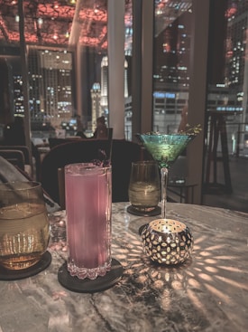 A dimly lit bar setting featuring a marble table with various drinks and a decorative candle holder casting intricate light patterns. A pink cocktail sits in a tall glass, accompanied by a green martini glass with a garnish. The background shows a window view of city buildings and lights against a nighttime sky.