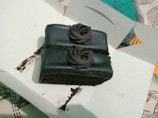 A rectangular piece of chocolate cake with dark chocolate frosting, situated inside a white cardboard box. The cake is topped with two rosettes made of chocolate cream. The background includes a colorful, patterned cloth.