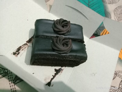 A rectangular piece of chocolate cake with dark chocolate frosting, situated inside a white cardboard box. The cake is topped with two rosettes made of chocolate cream. The background includes a colorful, patterned cloth.