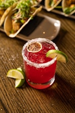 A vibrant red cocktail garnished with a slice of lime and a dehydrated orange slice sits on a wooden table. The rim of the glass is salted, adding a touch of elegance and contrast to the beverage. The background features a plate with tacos, suggesting a festive or dining setting.