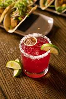A vibrant red cocktail garnished with a slice of lime and a dehydrated orange slice sits on a wooden table. The rim of the glass is salted, adding a touch of elegance and contrast to the beverage. The background features a plate with tacos, suggesting a festive or dining setting.