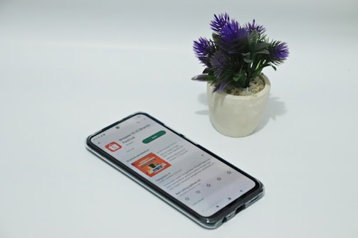 A smartphone displaying an app screen lays on a white surface next to a small potted plant with vibrant purple flowers.