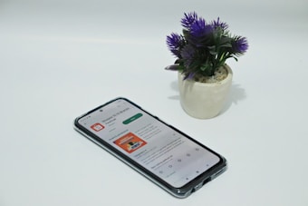 A smartphone displaying an app screen lays on a white surface next to a small potted plant with vibrant purple flowers.