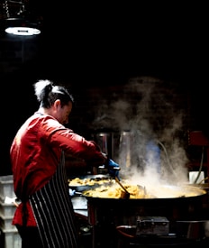 A person in a red shirt and striped apron is cooking in a large pan, surrounded by steam and lit by overhead lighting in a dim environment.