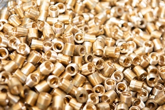 A large number of small, metallic threaded inserts are closely piled together. The inserts have a shiny gold appearance and are cylindrical in shape with hollow centers.