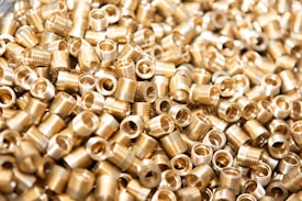 A large number of small, metallic threaded inserts are closely piled together. The inserts have a shiny gold appearance and are cylindrical in shape with hollow centers.