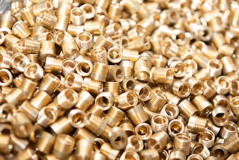 A large number of small, metallic threaded inserts are closely piled together. The inserts have a shiny gold appearance and are cylindrical in shape with hollow centers.