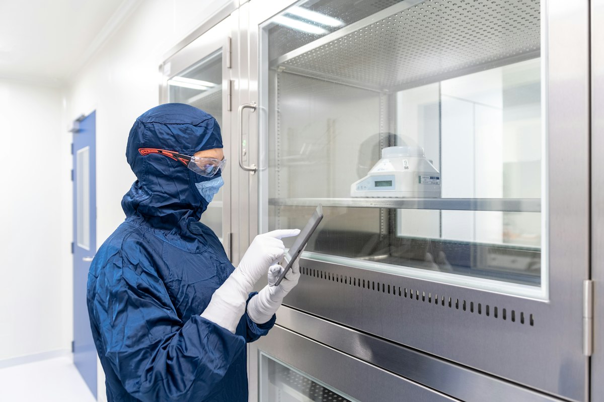 Laboratoriotyöntekijä sinisessä kasvomaskissa ja suojakäsineissä pitelee tablettitietokonetta.