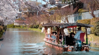 A traditional Japanese boat with several passengers leisurely cruises along a narrow canal lined with cherry blossom trees. The boat features rustic wooden construction and Japanese lanterns, with passengers enjoying the scenic view.