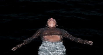 A person is floating on their back in dark water, with their head tilted upward and arms outstretched. The stillness of the water contrasts with the submerged parts of their body, creating a sense of calm and introspection.