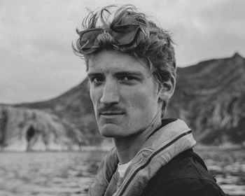 A black and white portrait of a person with wavy hair and sunglasses perched on the head. The background shows a rugged landscape with hills and water, suggesting an outdoor setting.