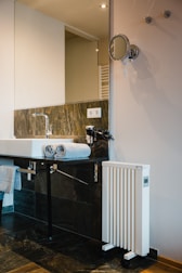 A modern bathroom setting featuring a white sink with a chrome faucet. Two neatly folded gray towels rest on the edge of the sink. The background includes a large rectangular mirror and a sleek wall-mounted makeup mirror. A white, vertical heater with wheels is positioned nearby. The wall is tastefully tiled with dark gray and brown tiles, complemented by a wooden floor.
