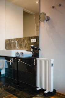 A modern bathroom setting featuring a white sink with a chrome faucet. Two neatly folded gray towels rest on the edge of the sink. The background includes a large rectangular mirror and a sleek wall-mounted makeup mirror. A white, vertical heater with wheels is positioned nearby. The wall is tastefully tiled with dark gray and brown tiles, complemented by a wooden floor.
