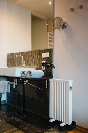 A modern bathroom setting featuring a white sink with a chrome faucet. Two neatly folded gray towels rest on the edge of the sink. The background includes a large rectangular mirror and a sleek wall-mounted makeup mirror. A white, vertical heater with wheels is positioned nearby. The wall is tastefully tiled with dark gray and brown tiles, complemented by a wooden floor.