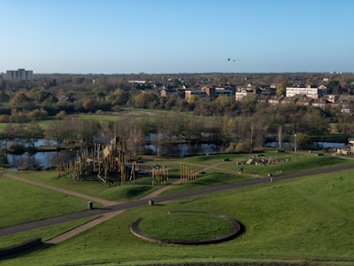 Aerial view of a park area with well-maintained green lawns, several pathways, and a central circular structure made of stone or earth. There is a wooden playground structure with slides and climbing areas. A small lake with surrounding trees and vegetation is visible in the background. Further back, a residential area with multi-story buildings and houses is seen, bordered by a forested area. The sky is clear and blue, and two birds are flying above the park.