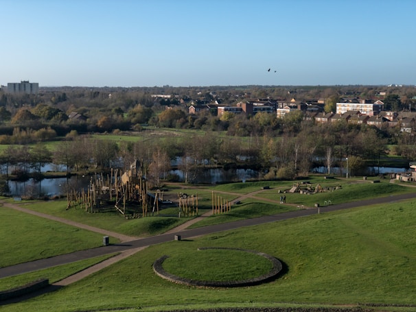Aerial view of a park area with well-maintained green lawns, several pathways, and a central circular structure made of stone or earth. There is a wooden playground structure with slides and climbing areas. A small lake with surrounding trees and vegetation is visible in the background. Further back, a residential area with multi-story buildings and houses is seen, bordered by a forested area. The sky is clear and blue, and two birds are flying above the park.