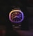 A sophisticated wristwatch with a metallic strap, illuminated by colorful lighting. The watch face displays Roman numerals and has a polished finish, adding to its elegance. It features a small window for the day and date on the right side. The overall appearance is sleek and luxurious.