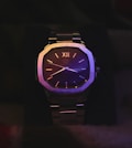 A sophisticated wristwatch with a metallic strap, illuminated by colorful lighting. The watch face displays Roman numerals and has a polished finish, adding to its elegance. It features a small window for the day and date on the right side. The overall appearance is sleek and luxurious.