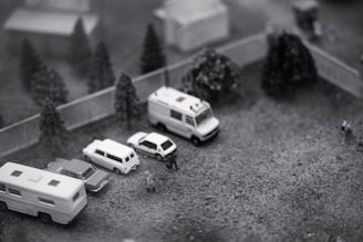 Several miniature vehicles, including vans and cars, are parked on a small gravel lot surrounded by trees and a fence. Small model figures are seen interacting around the vehicles.