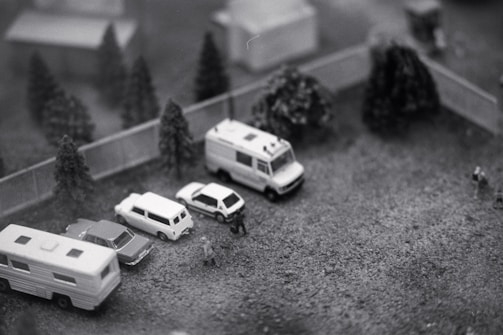 Several miniature vehicles, including vans and cars, are parked on a small gravel lot surrounded by trees and a fence. Small model figures are seen interacting around the vehicles.