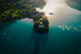 An aerial view of a serene lake surrounded by lush green hills and small villages. In the middle of the lake, there is a small island with a large house, surrounded by trees. Several small boats are scattered in the water, and a ferry can be seen making its way across the lake, creating a gentle wake. The sun casts a warm glow over the landscape, reflecting off the water's surface.
