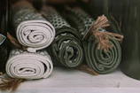 Four neatly rolled mats in shades of green and beige, each tied with a piece of twine, are arranged side by side on a shelf. The texture and patterns on the mats suggest they are woven or knitted.