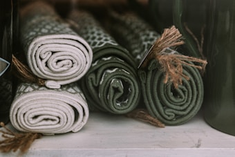 Four neatly rolled mats in shades of green and beige, each tied with a piece of twine, are arranged side by side on a shelf. The texture and patterns on the mats suggest they are woven or knitted.
