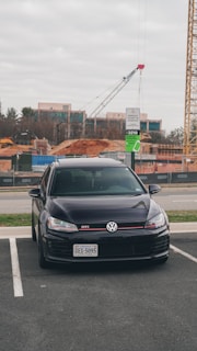 A black Volkswagen GTI is parked in an outdoor lot. In the background, there is a construction site with cranes, dirt piles, and unfinished buildings. A parking sign indicates reserved spaces, and the sky is overcast.