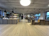 A spacious, modern interior features a wine bar with a large wooden counter and shelves displaying an assortment of wine bottles and products. Industrial-style pendant lights hang from the ceiling, and there are tables and high stools for seating. The room has wide windows, allowing plenty of natural light.