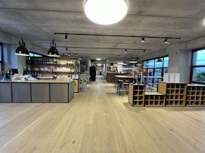 A spacious, modern interior features a wine bar with a large wooden counter and shelves displaying an assortment of wine bottles and products. Industrial-style pendant lights hang from the ceiling, and there are tables and high stools for seating. The room has wide windows, allowing plenty of natural light.