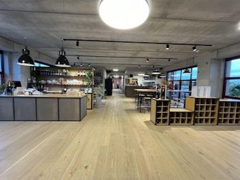 A spacious, modern interior features a wine bar with a large wooden counter and shelves displaying an assortment of wine bottles and products. Industrial-style pendant lights hang from the ceiling, and there are tables and high stools for seating. The room has wide windows, allowing plenty of natural light.