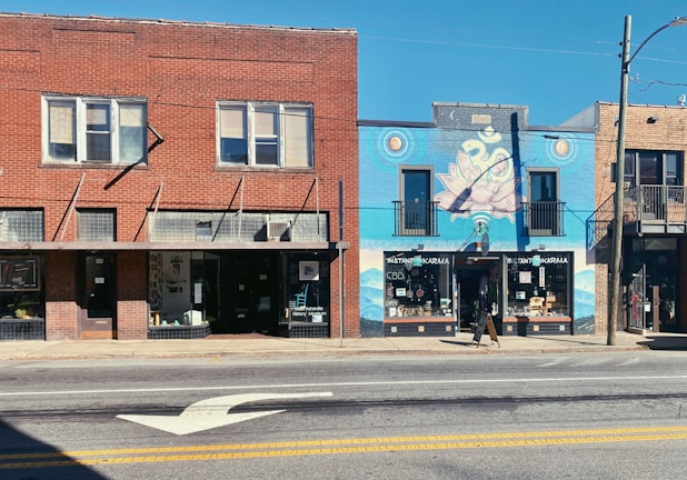 A street view of two adjacent buildings with distinct facades. The left building is red brick with multiple windows and a history museum sign. The right building features a vibrant blue mural incorporating spiritual symbols, including a lotus and an 'Om' symbol. There is a storefront named 'Instant Karma' beneath the mural.