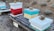 Several colorful beehives are placed on wooden pallets in an outdoor setting. The beehives are painted in bright red, teal, yellow, and white colors, each with a large stone on top. The area is surrounded by dry grass and a warning sign about an electric fence is visible. The environment suggests a beekeeping operation in a rural setting.