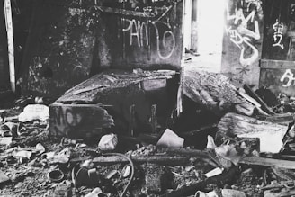 A dilapidated, abandoned room filled with debris and broken wooden structures. Graffiti covers the walls, and remnants of old, discarded items are scattered across the dirty floor.
