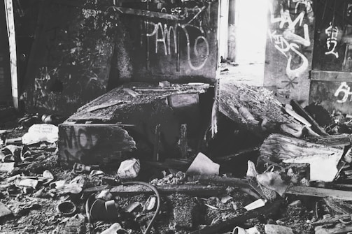 A dilapidated, abandoned room filled with debris and broken wooden structures. Graffiti covers the walls, and remnants of old, discarded items are scattered across the dirty floor.