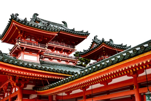 Traditional Japanese architecture with ornate, tiered roofs featuring intricate woodwork. The roof tiles are dark grey, contrasting with the vibrant red-orange and white of the building. The design is elaborate, indicating cultural and historical significance.