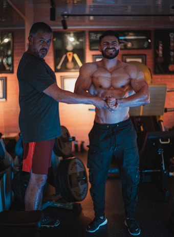 Two men are posing in a gym setting. One is muscular and shirtless, showcasing his physique, while the other is standing beside him with a proud expression. The background includes gym equipment and framed awards on the wall, suggesting a bodybuilding or fitness theme.