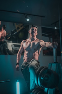 A muscular man with defined abs is standing confidently in a gym, his arm resting on a piece of fitness equipment. The environment suggests a focused and intense workout session, with weights and exercise machines visible in the background. The lighting casts subtle shadows, highlighting the man's physique.